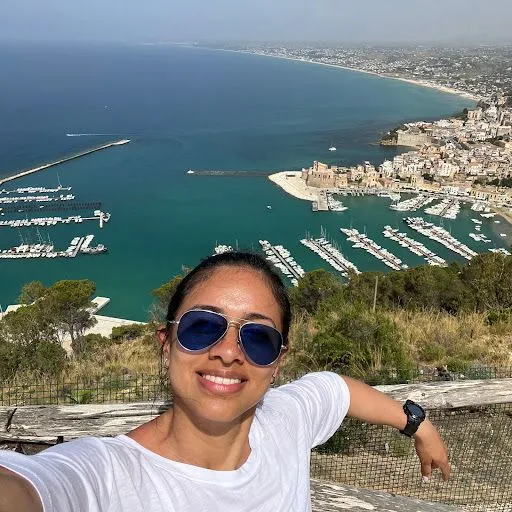 A woman wearing sunglasses is taking a selfie with a city in the background.